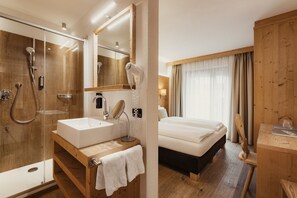 A hotel room with a wooden floor, a large bed, a wooden desk, a bathroom with a glass shower enclosure, a sink, and a mirror. - Diana Dolomites  (Badia)