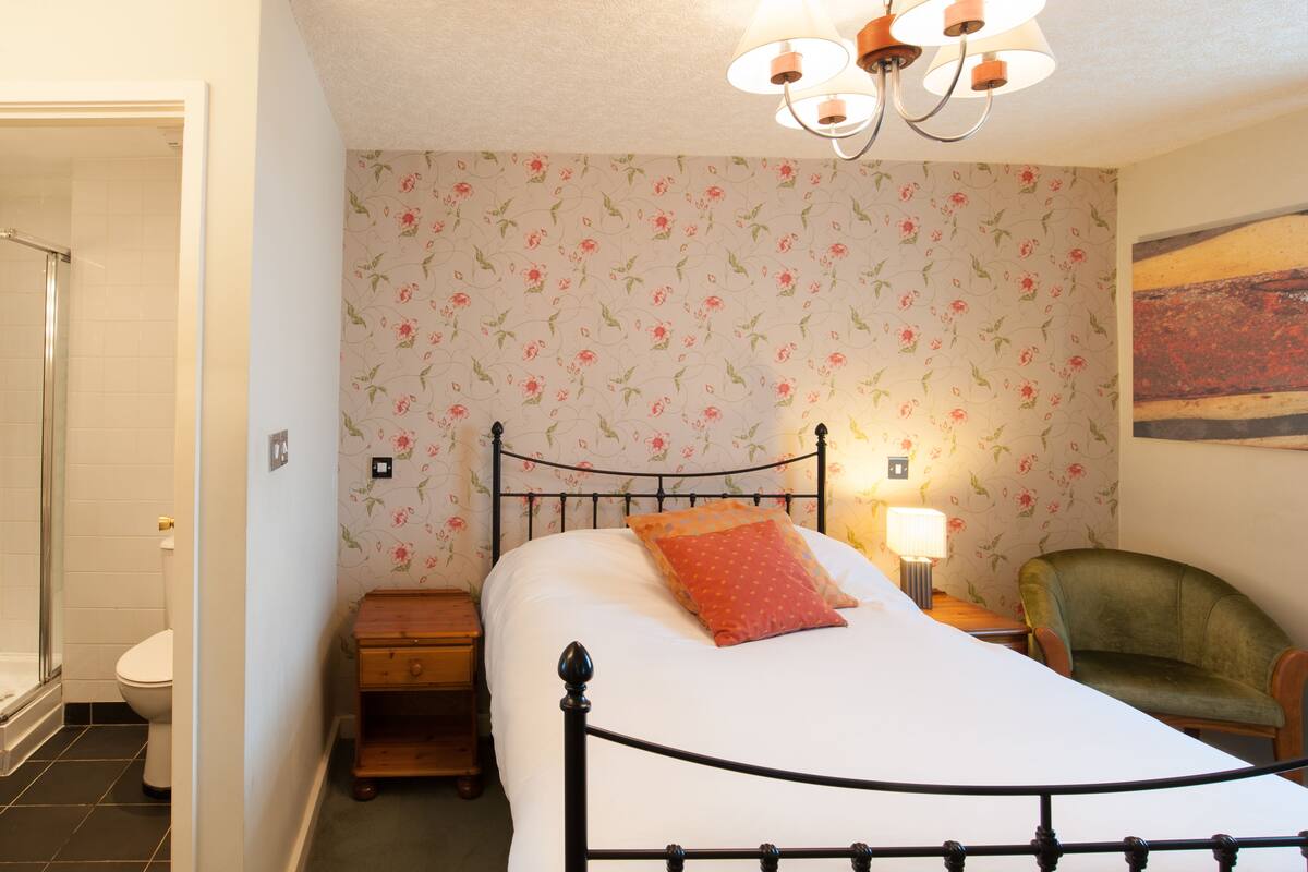 A bedroom with a bed, a nightstand, a chair, a lamp, and a shower.