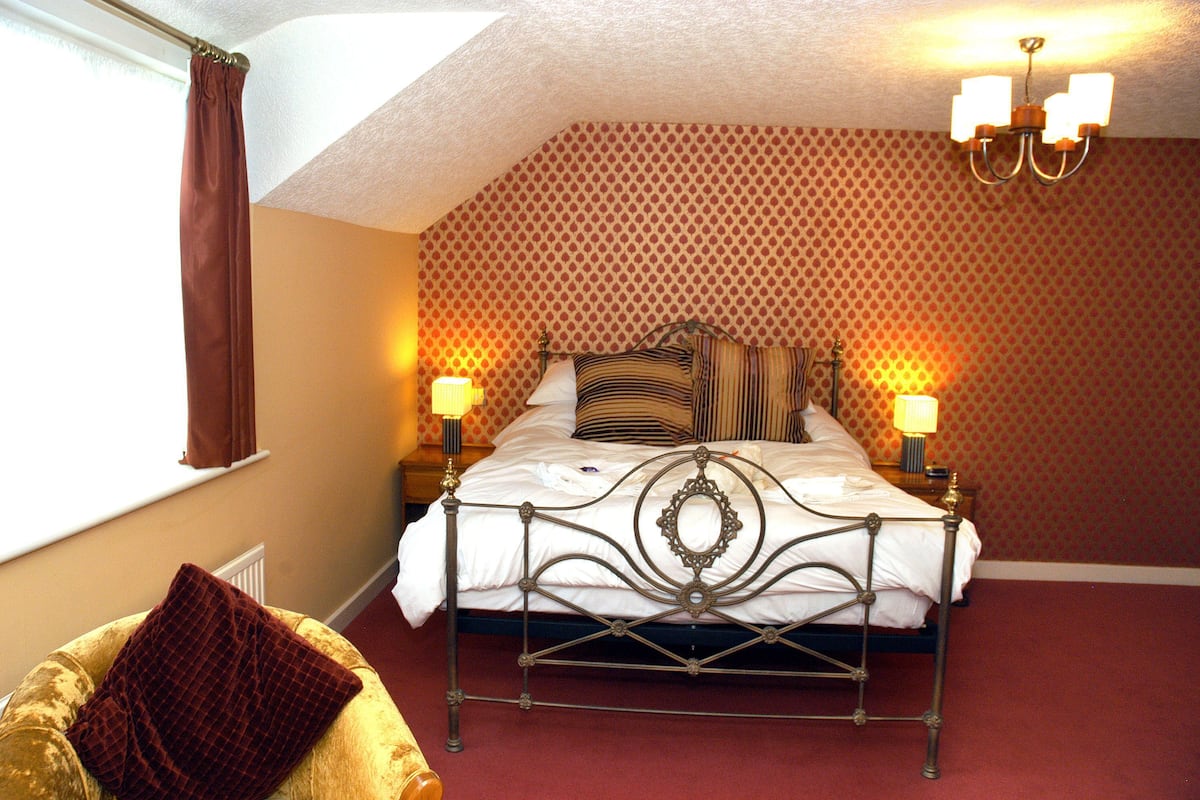 A bedroom with a metal bed frame, two bedside lamps, patterned wallpaper, and a window with a curtain.