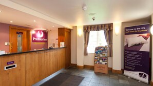 A hotel reception area with a wooden reception desk, a private door, and a welcome sign for business guests. - Premier Inn Manchester - Handforth (Wilmslow)
