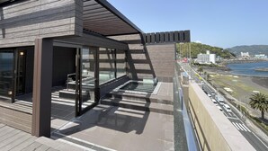 A modern building with large windows, a covered outdoor area, and a view of the sea and cityscape. - Kameya-Rakan (Ito)