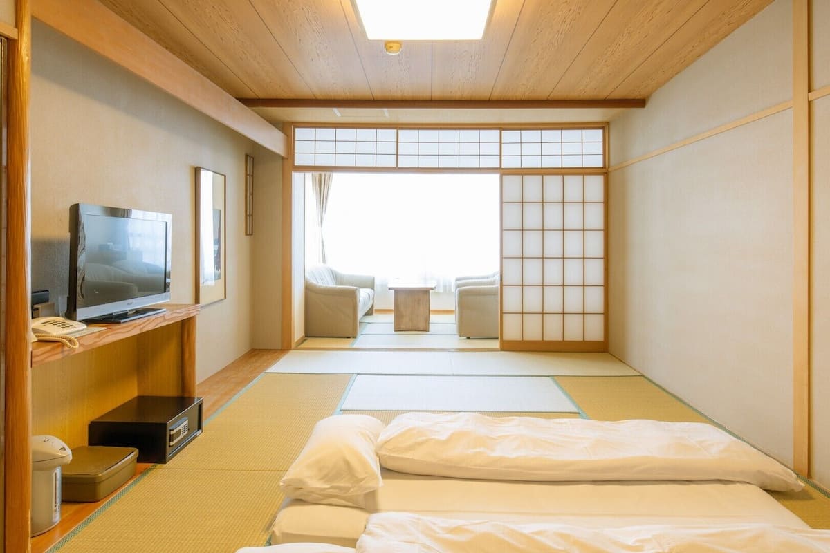 Une pièce de style japonais traditionnel, avec un sol recouvert de tatamis, une télévision et une porte coulissante donnant sur un espace salon.