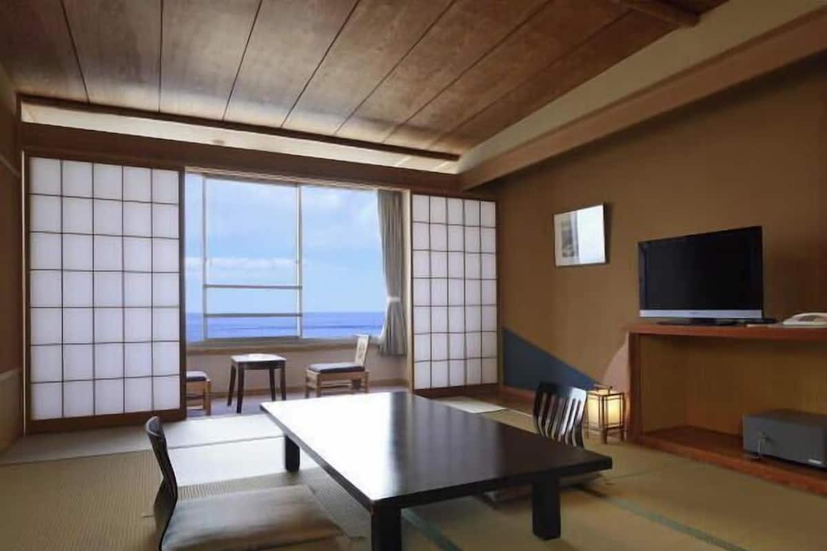 Une pièce de style japonais traditionnel, avec un sol recouvert de tatamis, une table basse, des chaises, un téléviseur et une vue sur la mer.