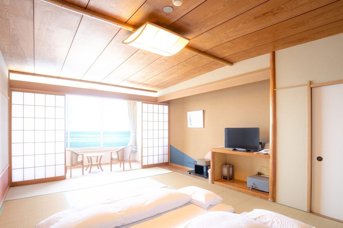 Une pièce avec un sol recouvert de tatamis, un lit, une télévision et une vue sur la mer.