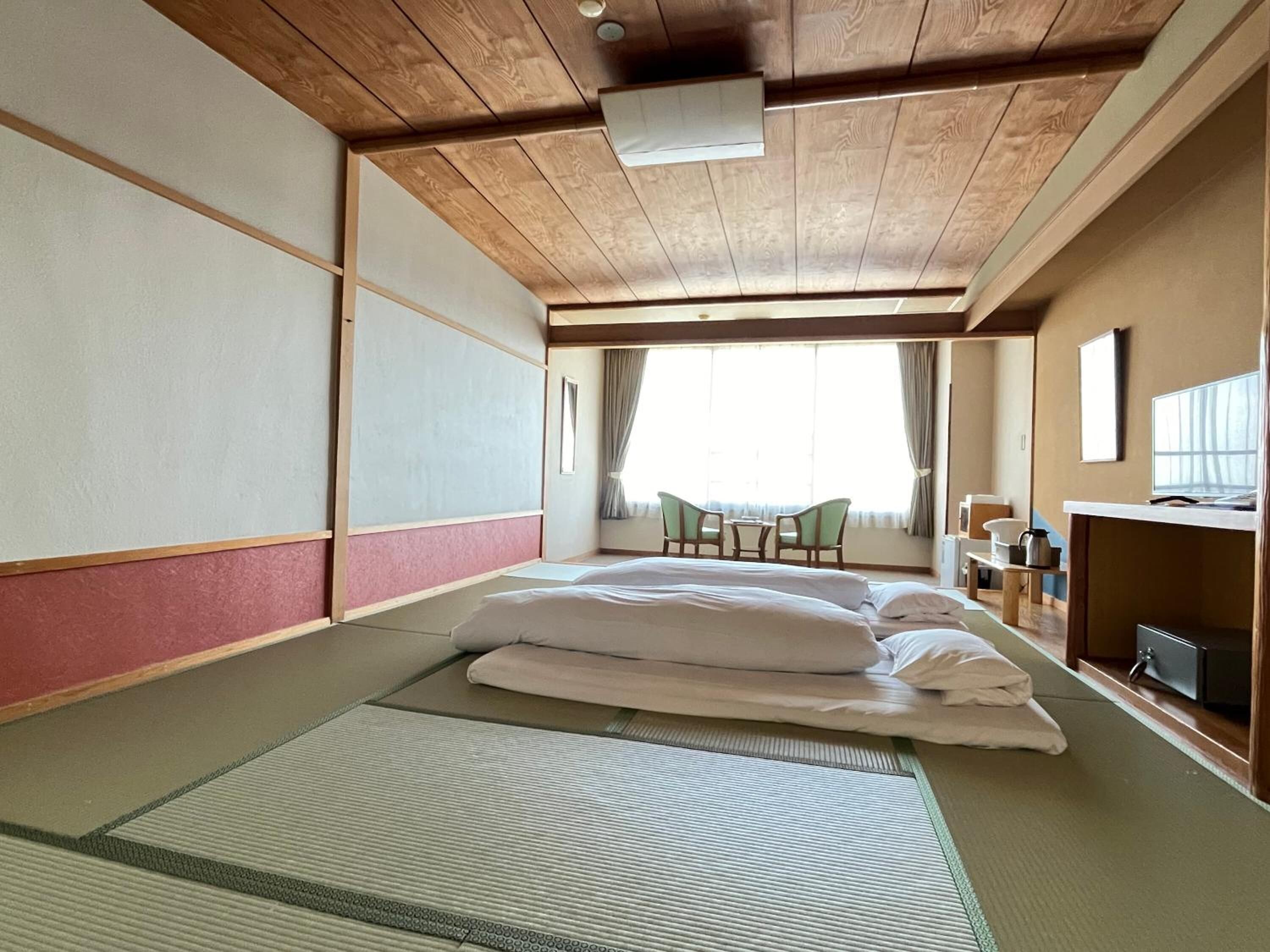 Um quarto japonês tradicional com tatame, uma cama baixa, uma escrivaninha e vista para o exterior.