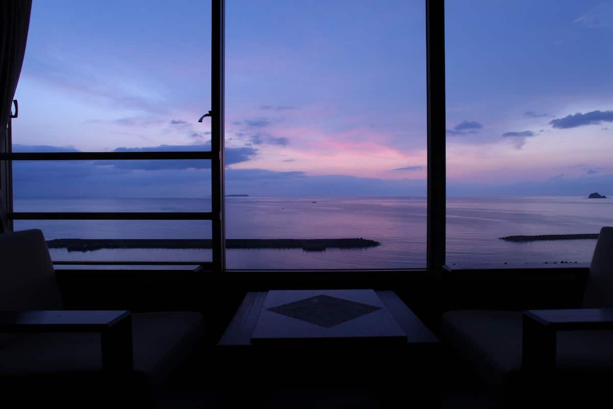 Une pièce dotée d’une grande fenêtre donnant sur une mer calme et sur une terre lointaine, avec une disposition simple comprenant deux chaises et une petite table.