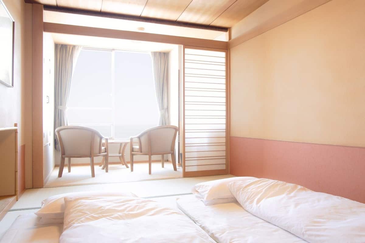 Une chambre d’hôtel avec un lit, deux chaises, une petite table et une vue par une fenêtre avec des rideaux.