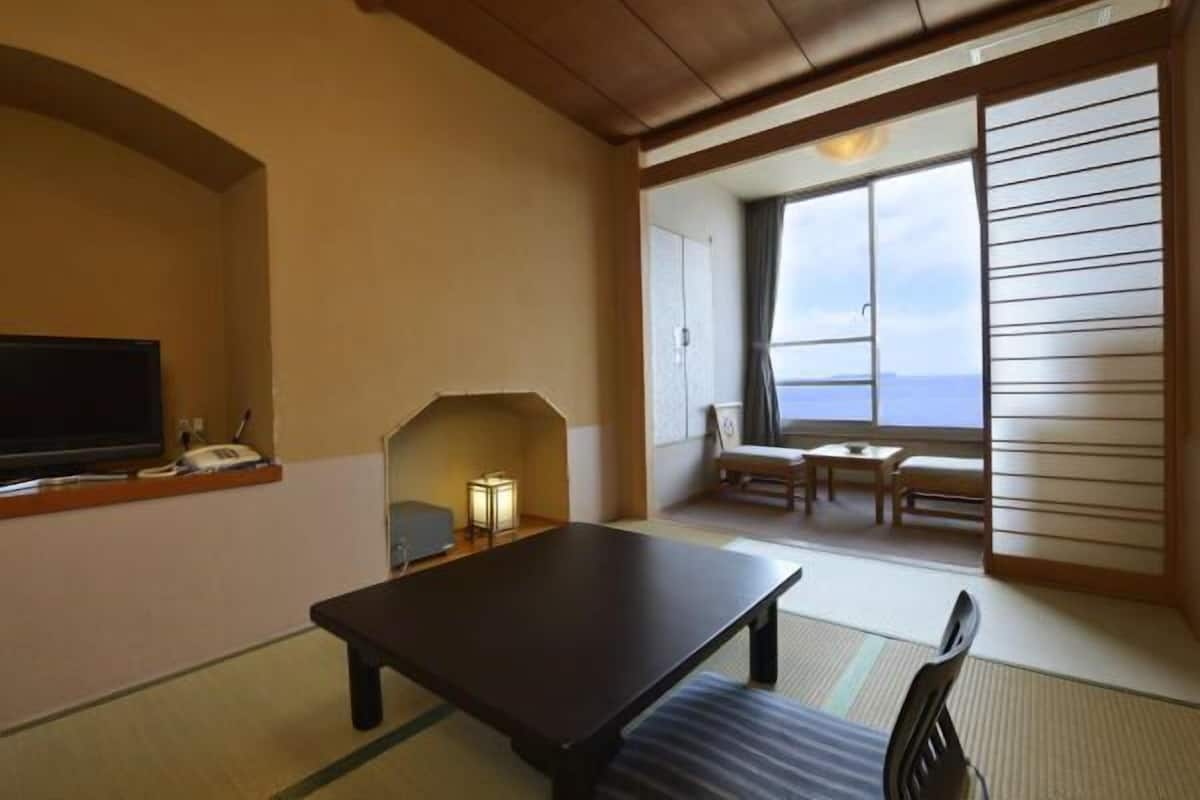 Une pièce dotée d’un sol recouvert de tatamis, d’une table basse, d’un téléviseur et offrant une vue sur la mer grâce à de grandes fenêtres.