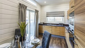 A modern kitchen with wooden cabinets, a dining area with a table set for four, and a large window with curtains. - Roydon Marina Village (Harlow)