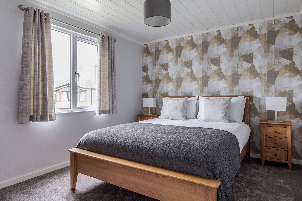 A bedroom with a wooden bed, two bedside tables with lamps, a window with curtains, and a geometric-patterned wall.