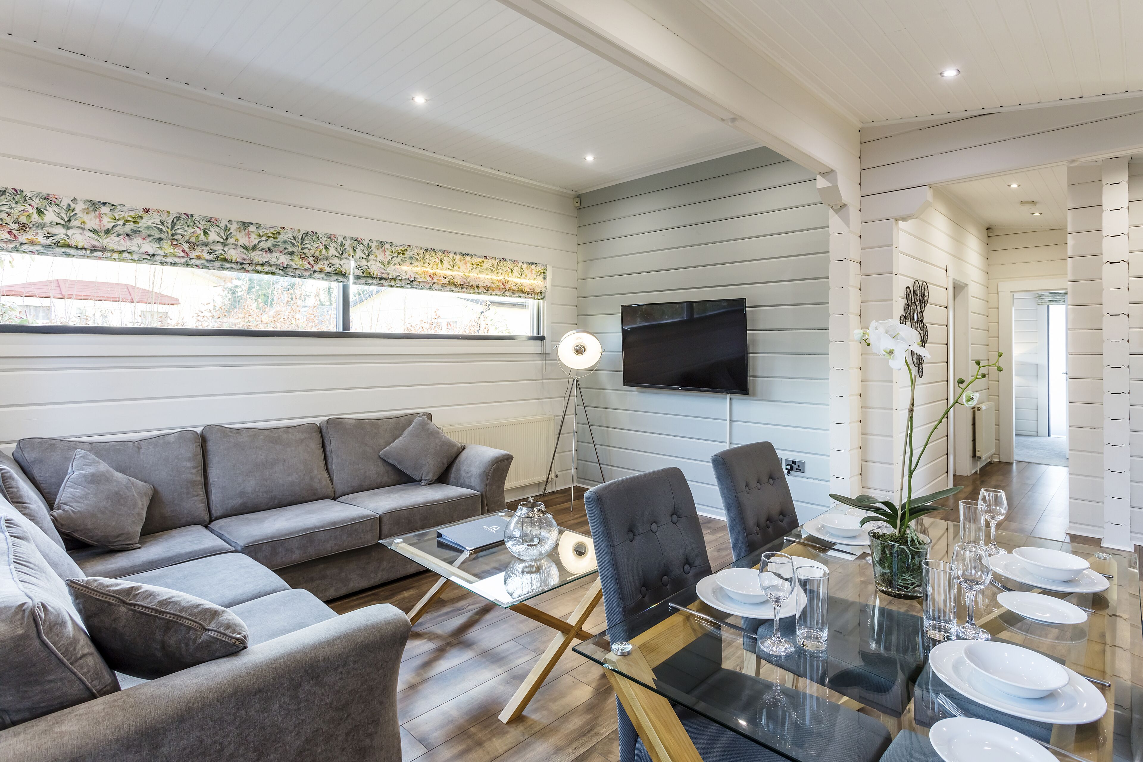 A modern living room with a grey sofa, a glass coffee table, and a dining area with a glass table set for six.