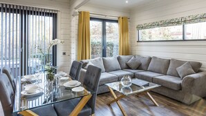 A modern living room with a glass dining table set for six, a grey sectional sofa, and a large window with yellow curtains. - Roydon Marina Village (Harlow)