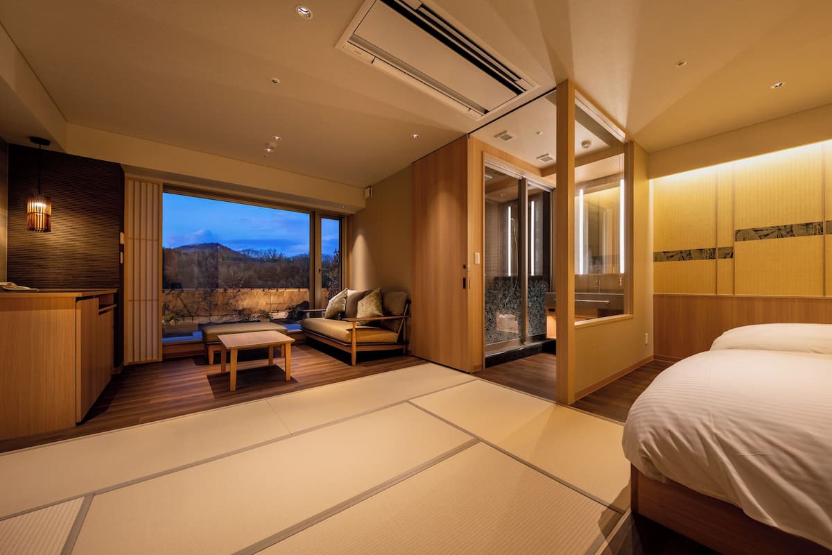 Une chambre d’hôtel comprenant un lit, un coin salon avec vue, une porte coulissante donnant sur un balcon et un comptoir d’accueil en bois.
