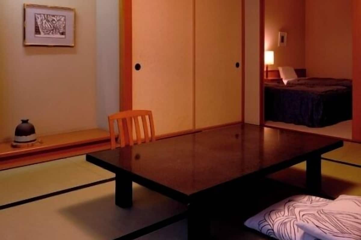 Une pièce de style japonais traditionnel, avec un sol recouvert de tatamis, une table basse, une chaise en bois, un lit recouvert d’une couverture sombre et un luminaire suspendu.