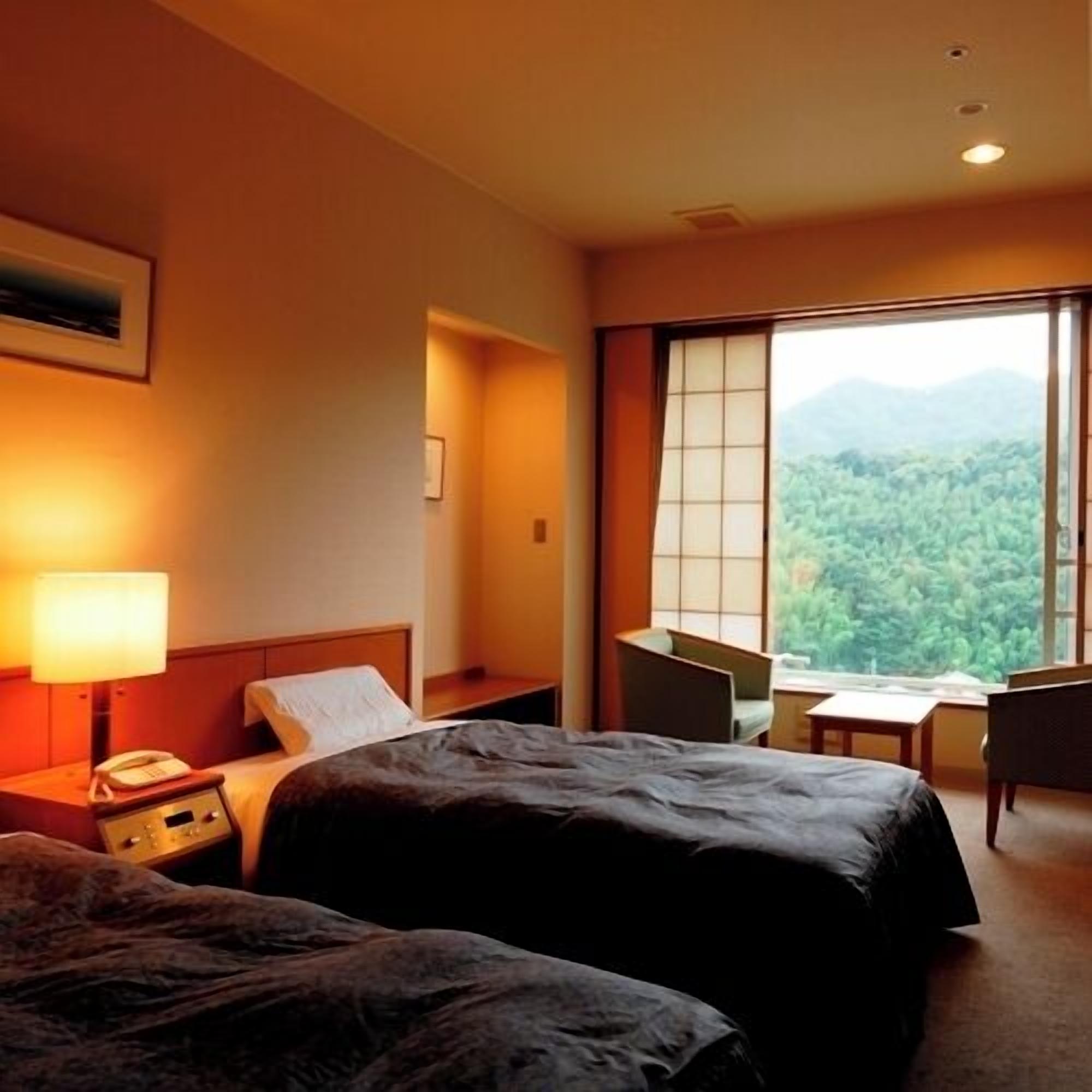 A hotel room with two beds, a desk, a chair, and a large window with a mountain view.