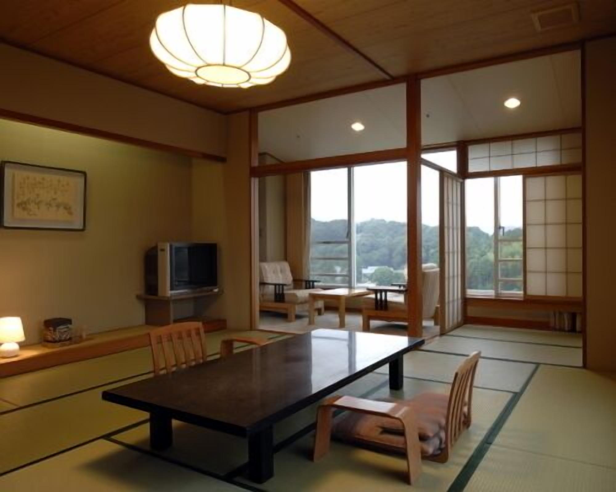 Ein traditioneller japanischer Raum mit Tatami-Boden, einem niedrigen Tisch, Stühlen, einem Fernseher und großen Fenstern mit Blick ins Freie.