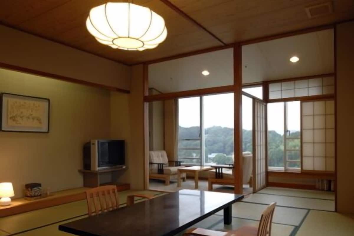 Une pièce de style japonais traditionnel, avec un sol recouvert de tatamis, une table basse, des chaises, un téléviseur et de grandes fenêtres donnant sur l’extérieur.
