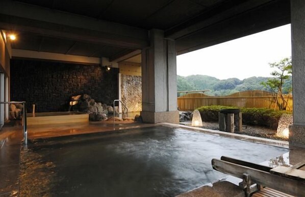 A stone-tiled indoor pool area with a stone wall and a view of a wooden fence and greenery. - Izu Nagaoka Hotel Tenbo (Izunokuni)