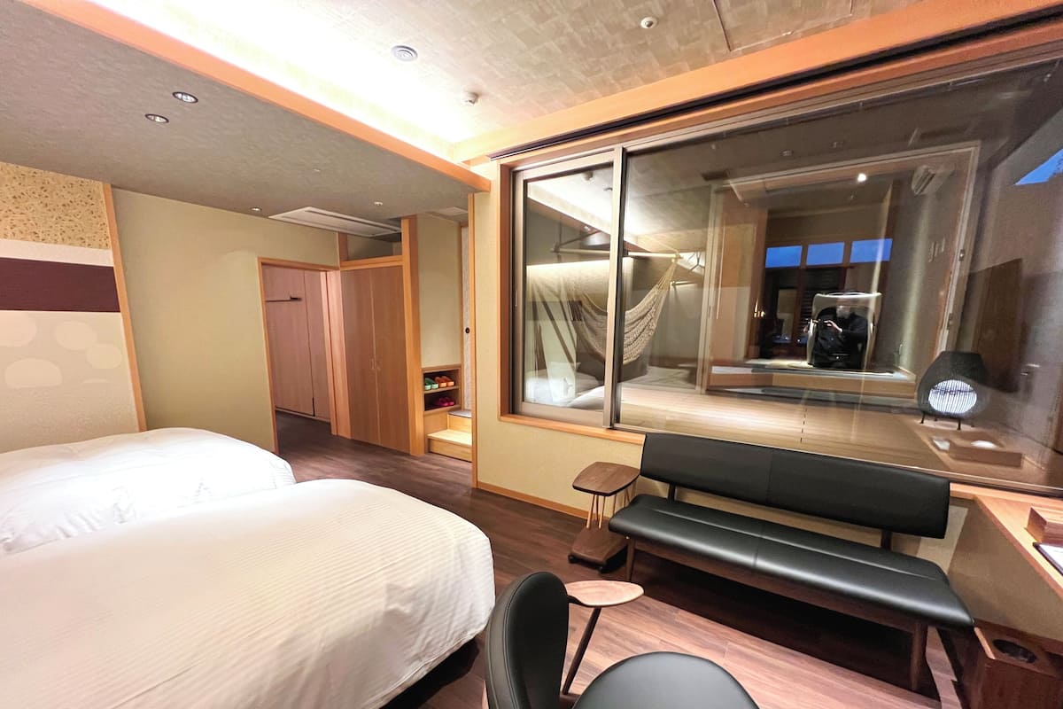 Une chambre d’hôtel comprenant un lit, une banquette, un bureau, une chaise, une armoire et une grande fenêtre donnant sur un paysage agréable.