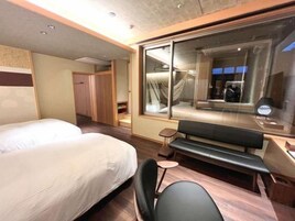 Habitación de hotel con dos camas, un banco, una mesita pequeña y un ventanal grande.