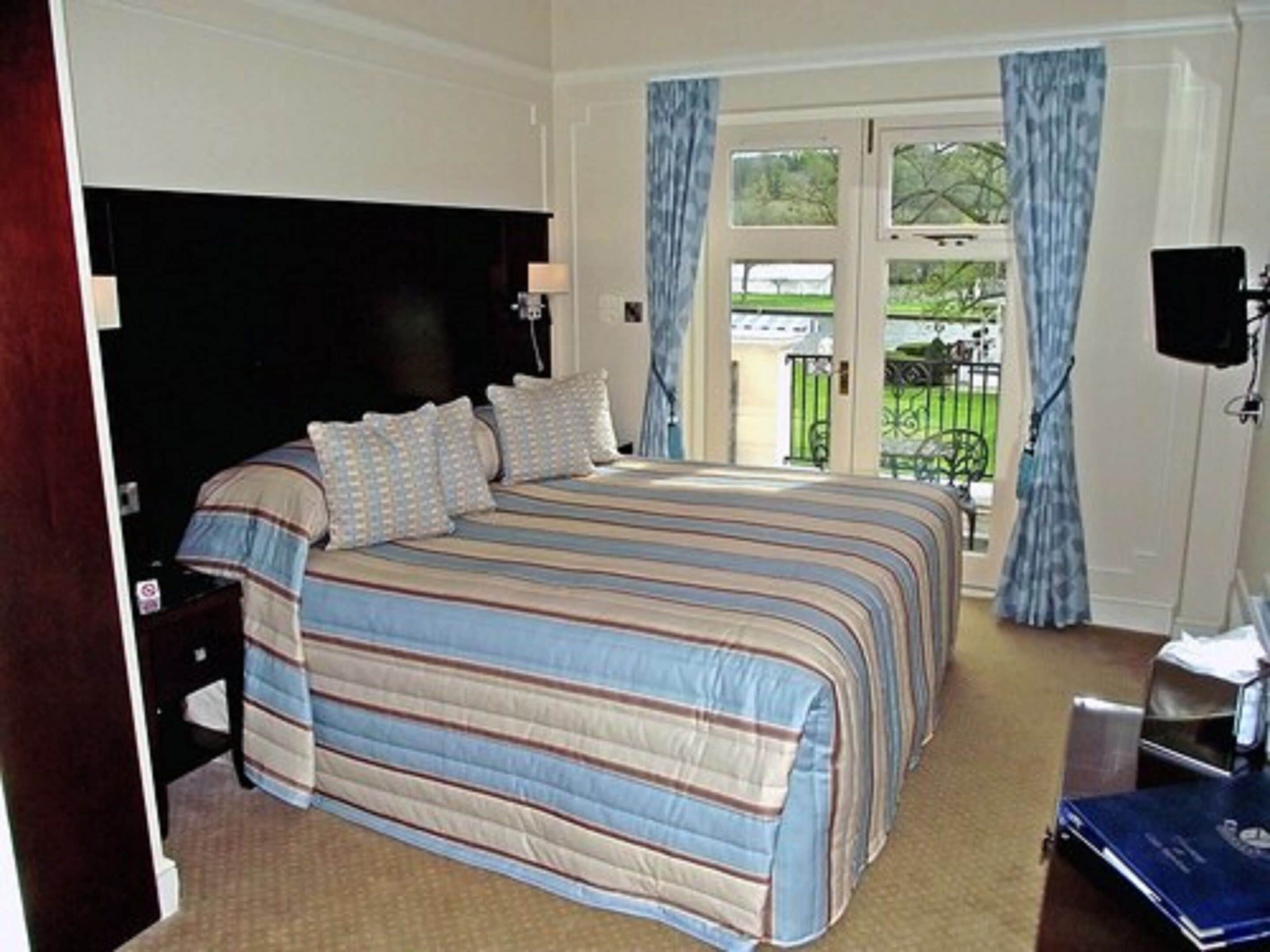 A bedroom with a large bed, a nightstand, a lamp, a window with curtains, and a view of greenery outside.