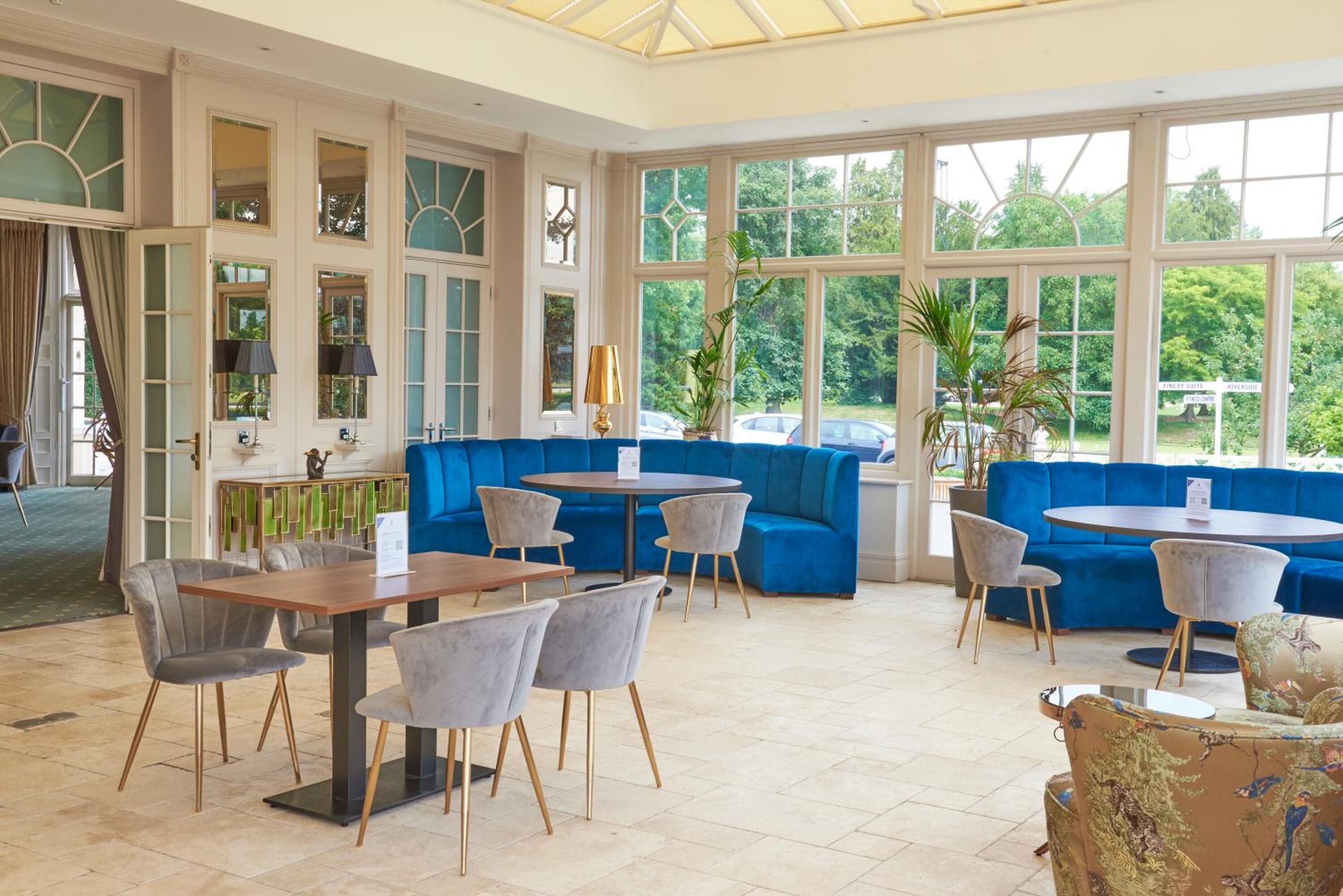 A spacious indoor area with blue seating, tables, and chairs, large windows, and a view of greenery outside.