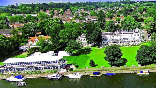 A riverside view of a town with a large white building, several smaller houses, and boats docked along the water. - Phyllis Court Members Club (Henley-on-Thames)