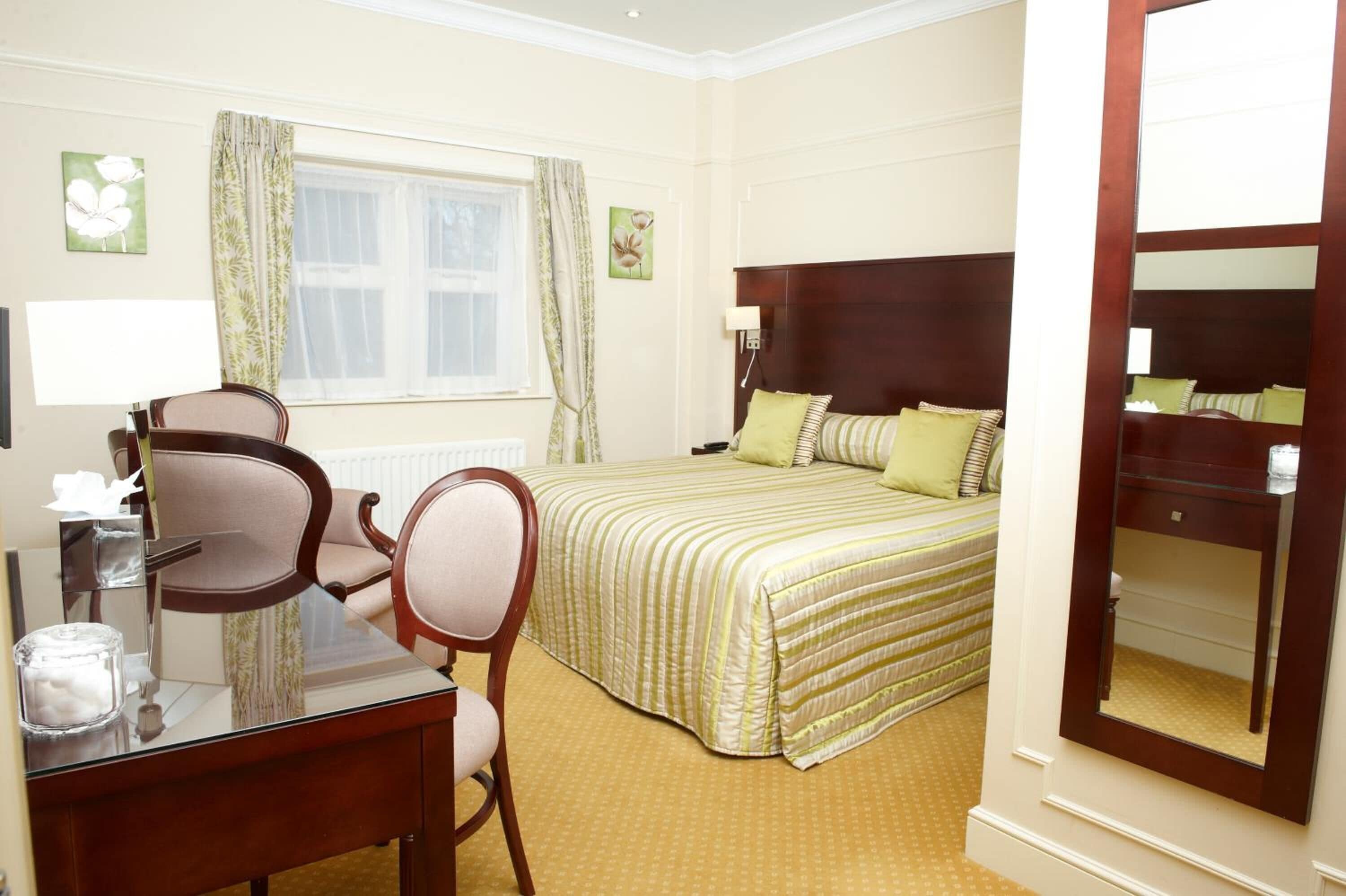 A hotel room with a bed, a desk with a chair, a window with curtains, and a mirror.