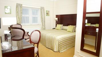 A hotel room with a bed, a desk with a chair, a window with curtains, and a mirror.