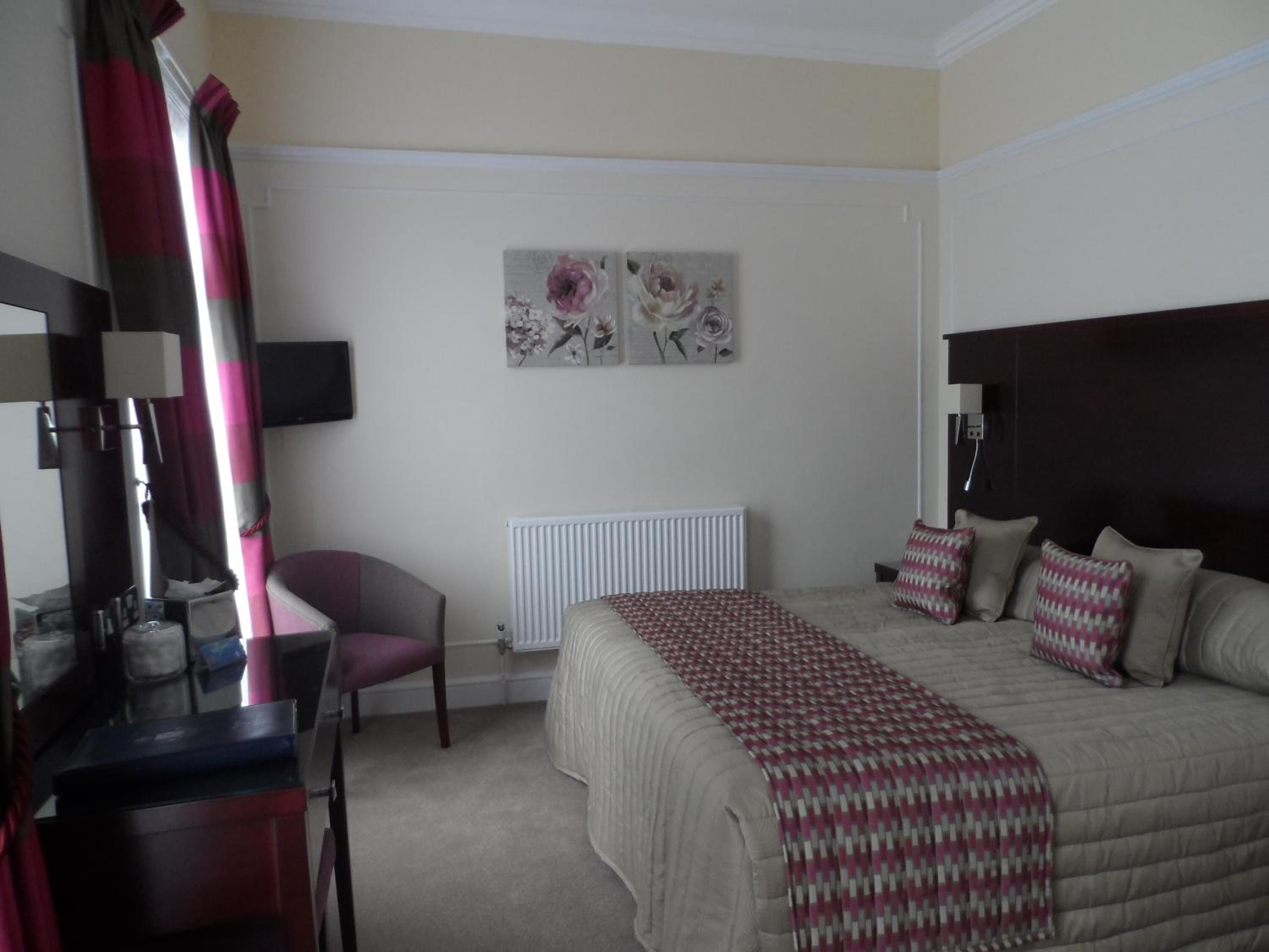 A hotel room with a bed, a desk, a chair, a radiator, and two wall-mounted artworks.