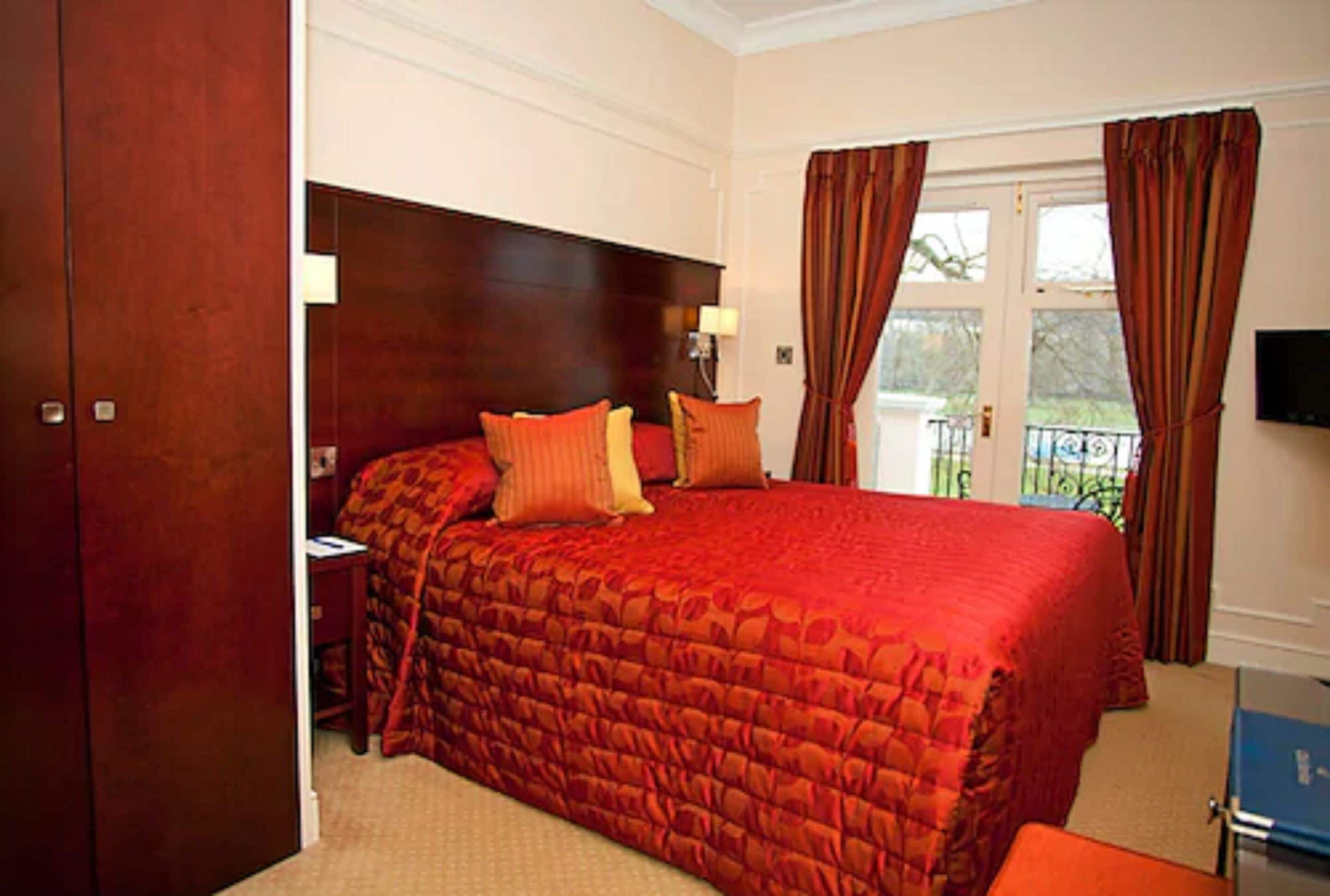 A bedroom with a large bed, a wooden wardrobe, a nightstand, and a view of the outdoors through a window with curtains.