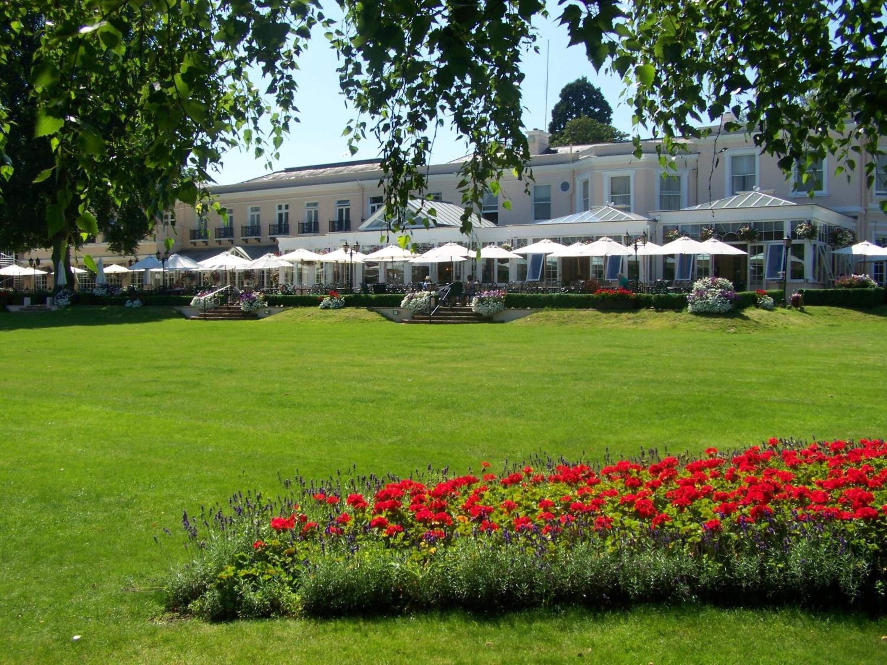 Un hotel con un amplio césped, zona de estar exterior con sombrillas y un parterre con flores rojas.