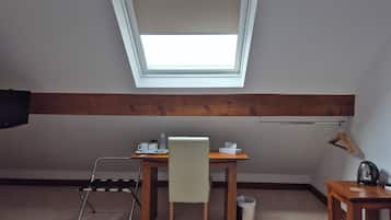 A small, well-lit room with a sloped ceiling, a wooden table with a chair, a small sofa, a bedside table with a kettle, and a window with a view.