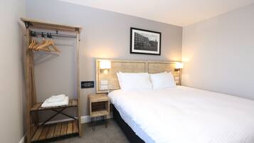 A hotel room with a large bed, a wooden wardrobe with hangers, a small bedside table with a lamp, and a framed picture on the wall.