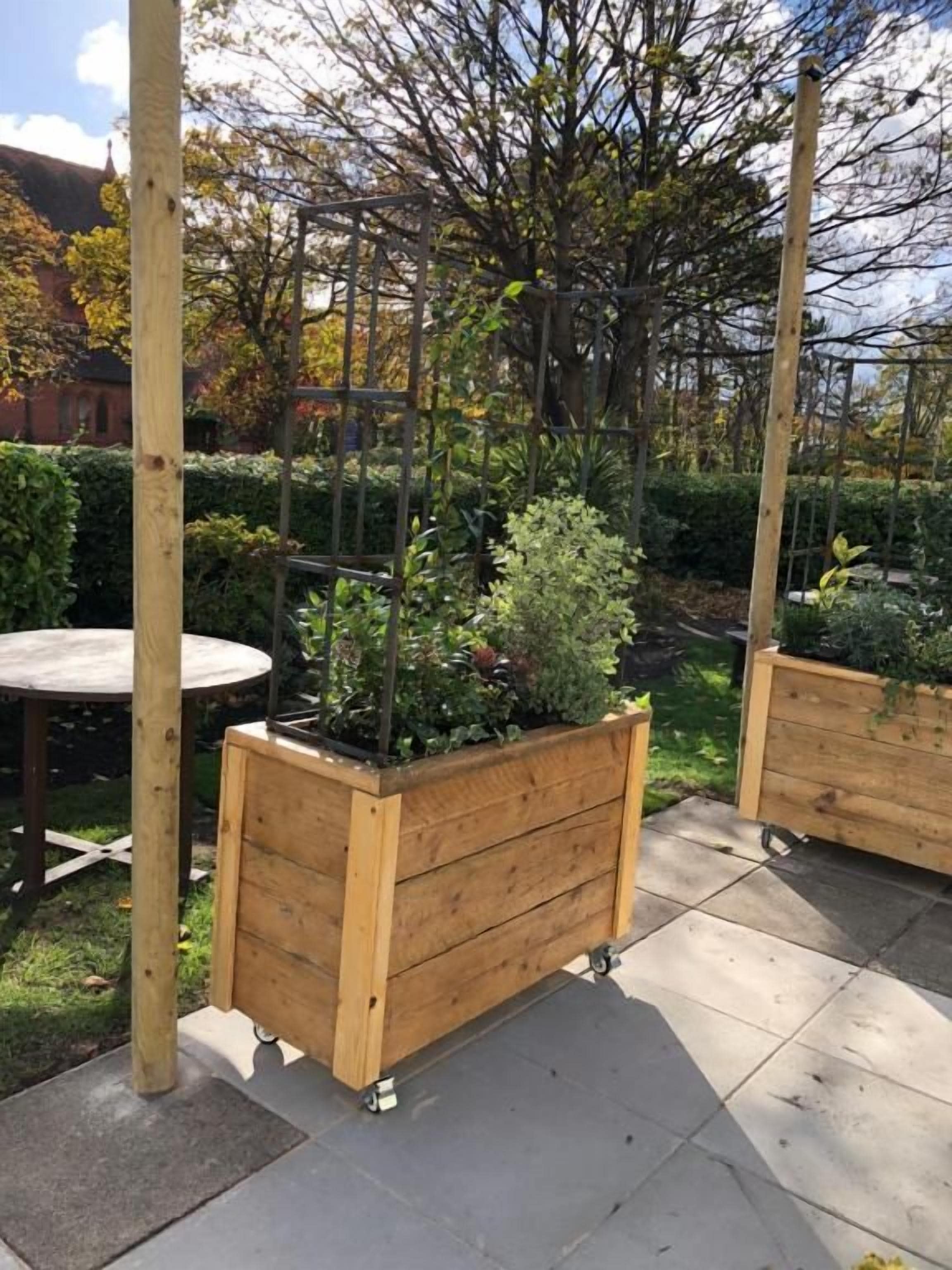 Un giardino con fioriere in legno su ruote, un pergolato in legno e un tavolo rotondo.