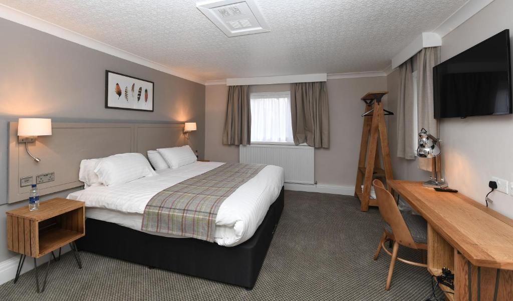 A hotel room with a bed, bedside table, lamp, desk, chair, TV, and window with curtains.