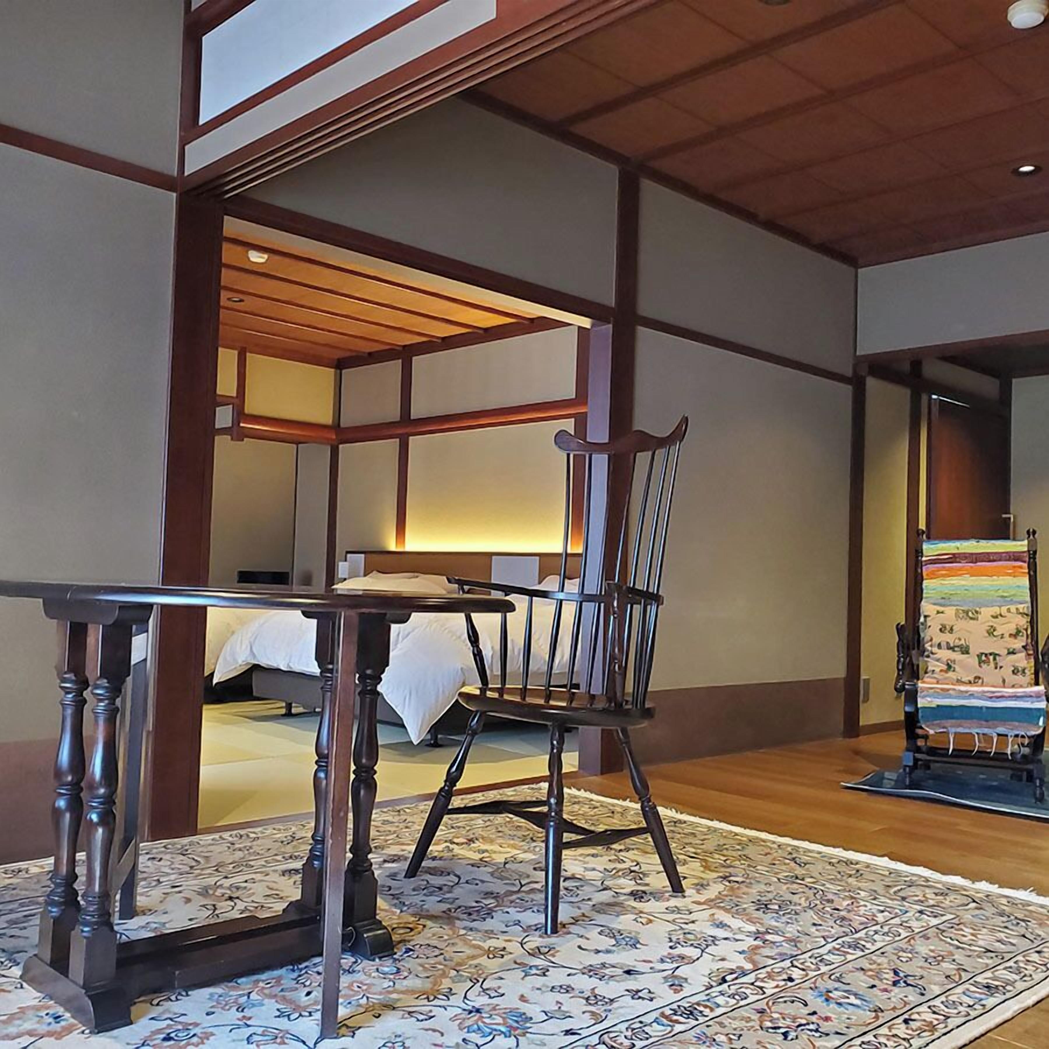 Een traditionele Japanse kamer met tatami vloerbedekking, een lage houten tafel, een stoel en een bed met een witte dekbedovertrek.