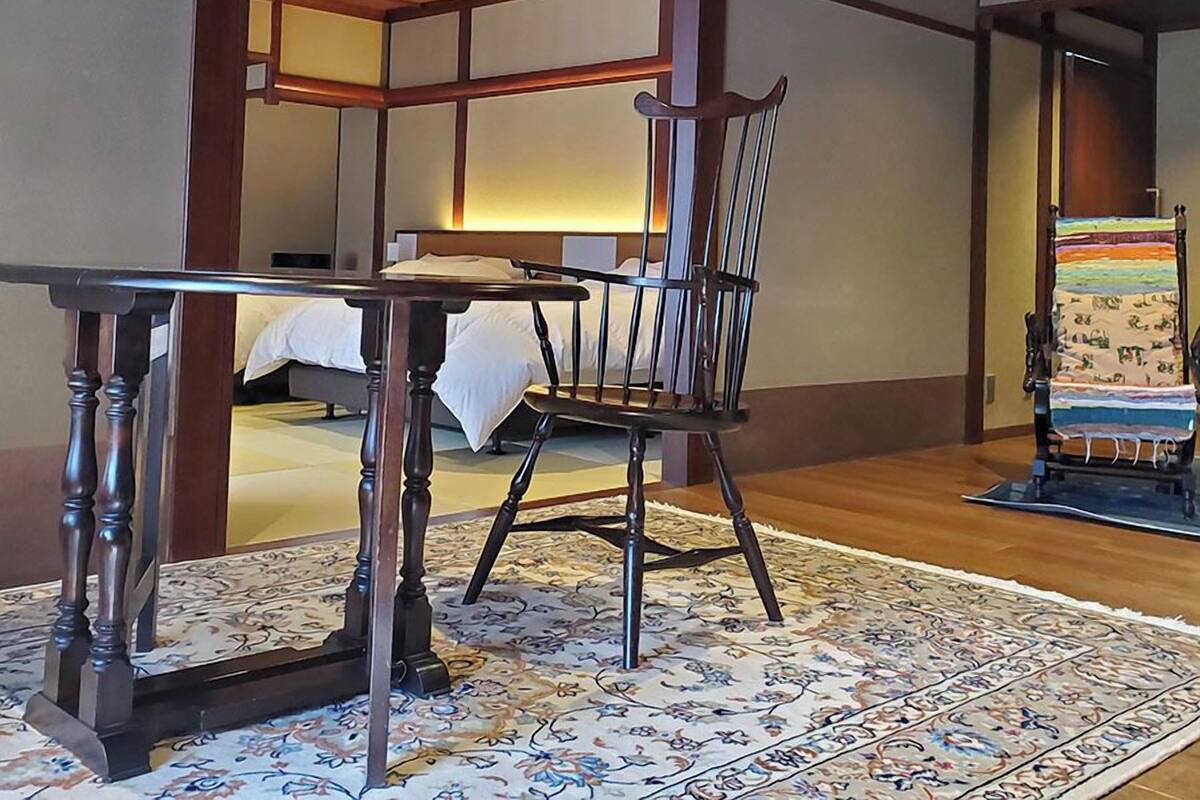 Une pièce de style japonais traditionnel, avec un sol recouvert de tatamis, une petite table en bois, une chaise et un lit recouvert d’une couverture blanche.
