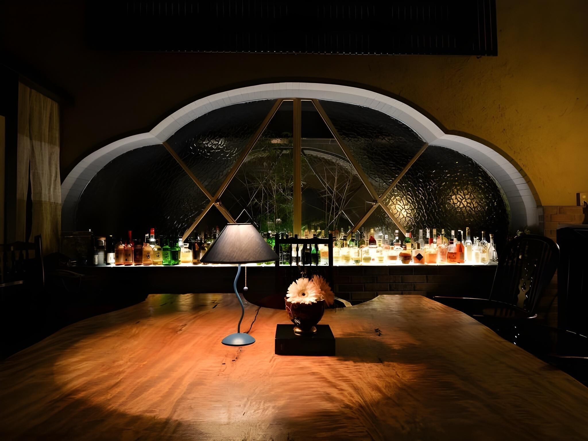 Zona de bar amb un gran finestral apuntat, una taula amb una làmpada i diverses ampolles.