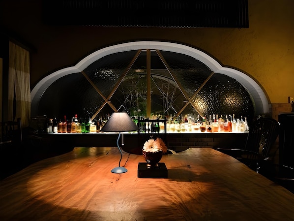 A bar area with a large arched window, a table with a lamp, and a variety of bottles.