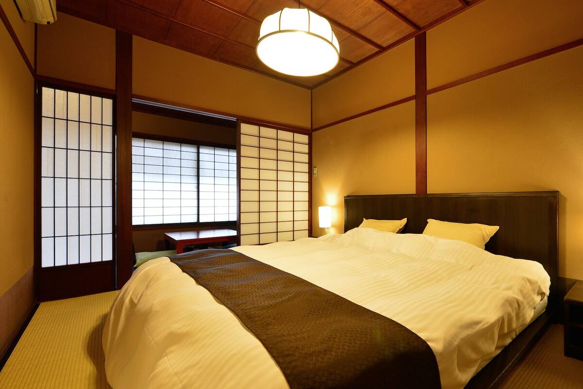 Une chambre de style japonais traditionnel, avec un sol recouvert de tatamis, un lit bas en bois, des paravents shoji et un luminaire suspendu.