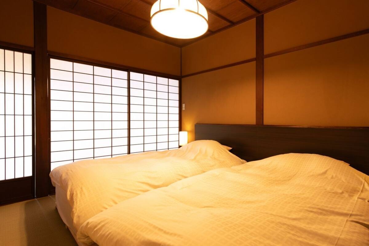 Une chambre de style japonais traditionnel, avec un sol recouvert de tatamis, une tête de lit en bois basse et une fenêtre à panneaux coulissants de type shoji.