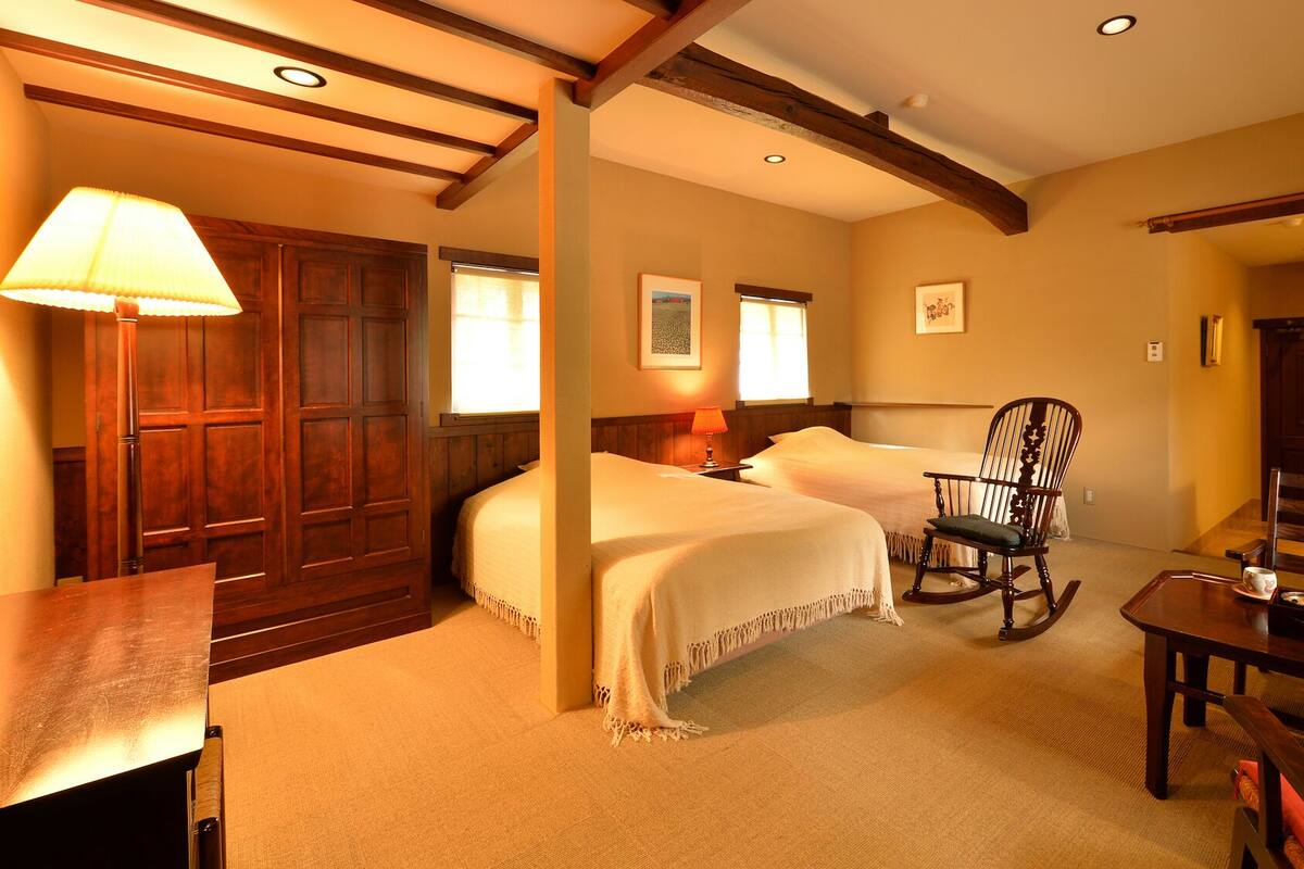 Une chambre d’hôtel avec deux lits, un fauteuil à bascule en bois, un bureau avec une télévision et une lampe.