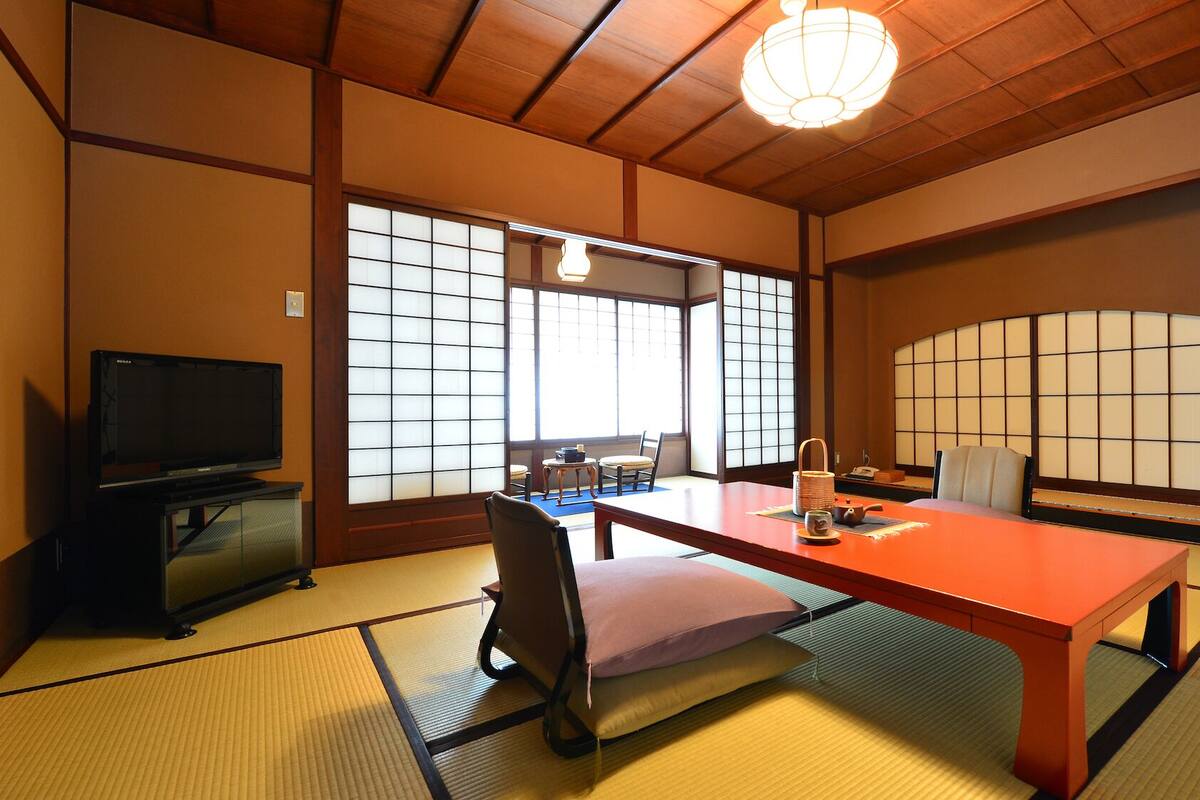 Une pièce de style japonais traditionnel, avec un sol recouvert de tatamis, un téléviseur, une table avec un coussin et des portes coulissantes dotées de panneaux shoji.