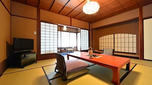 Ein traditioneller japanischer Raum mit Tatami-Boden, einem Fernseher, einem Tisch mit Kissen und einer hängenden Leuchte.