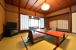 A traditional Japanese room with tatami flooring, a television, a table with a cushion, and a hanging light fixture.
