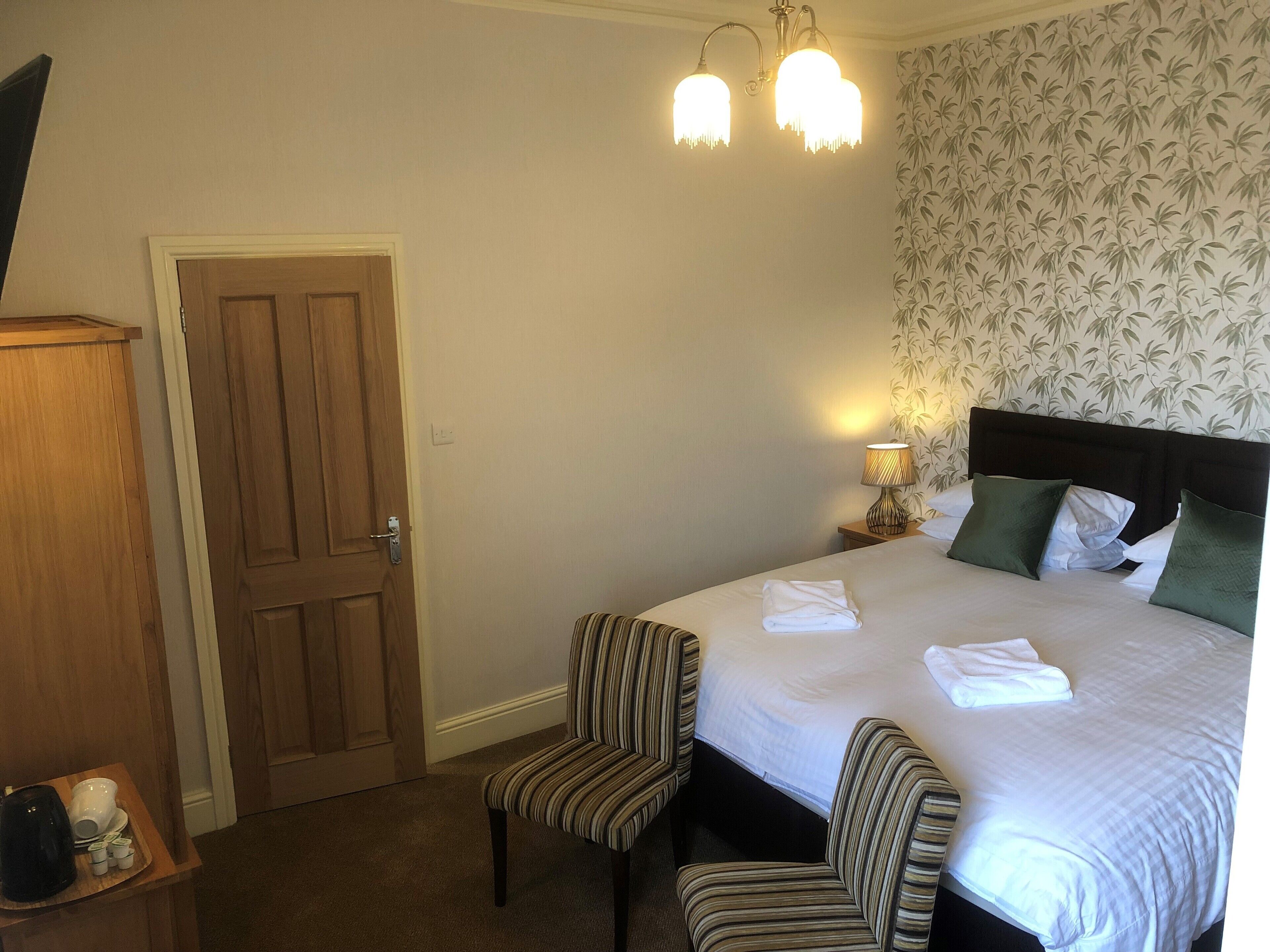 Een hotelkamer met een bed, twee gestreepte fauteuils, een houten garderobe en een nachtkastje met een lamp.