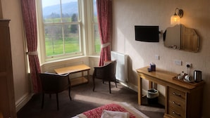 A room with a large window, a desk, a chair, a radiator, a wooden cabinet, and a television mounted on the wall.