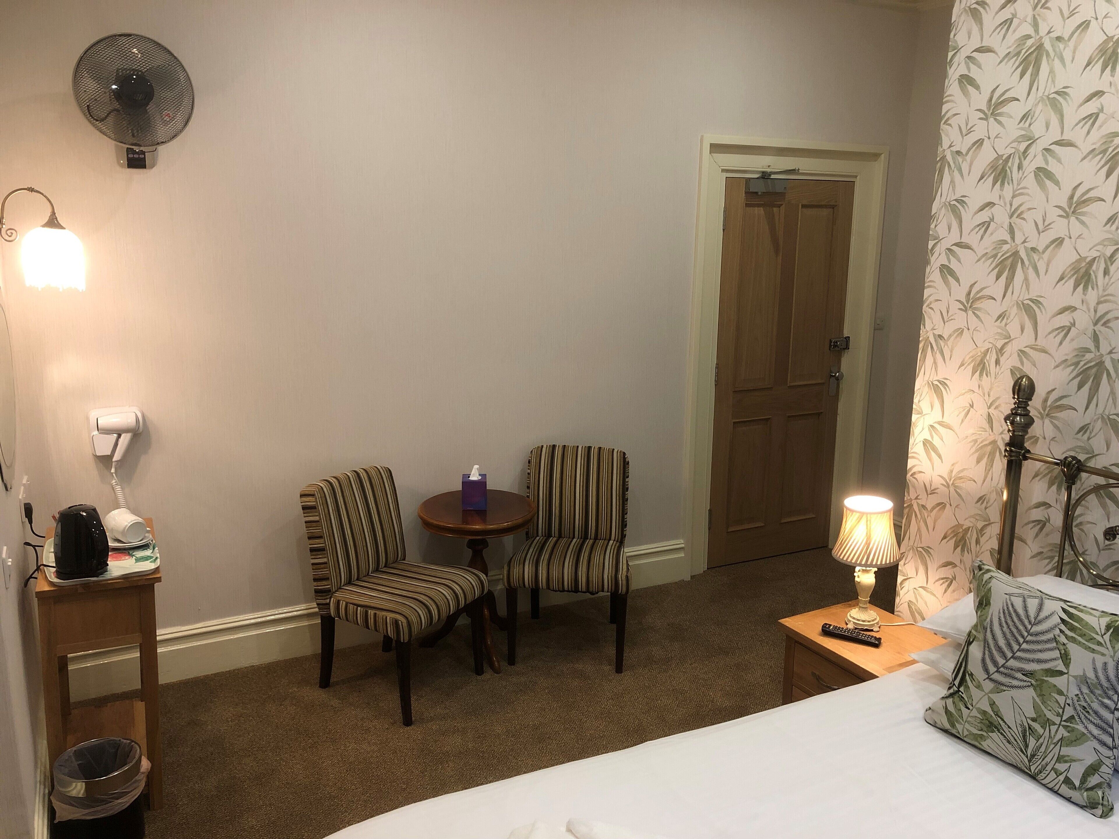 A hotel room with a bed, two striped chairs, a small table, a fan, a lamp, and a door.