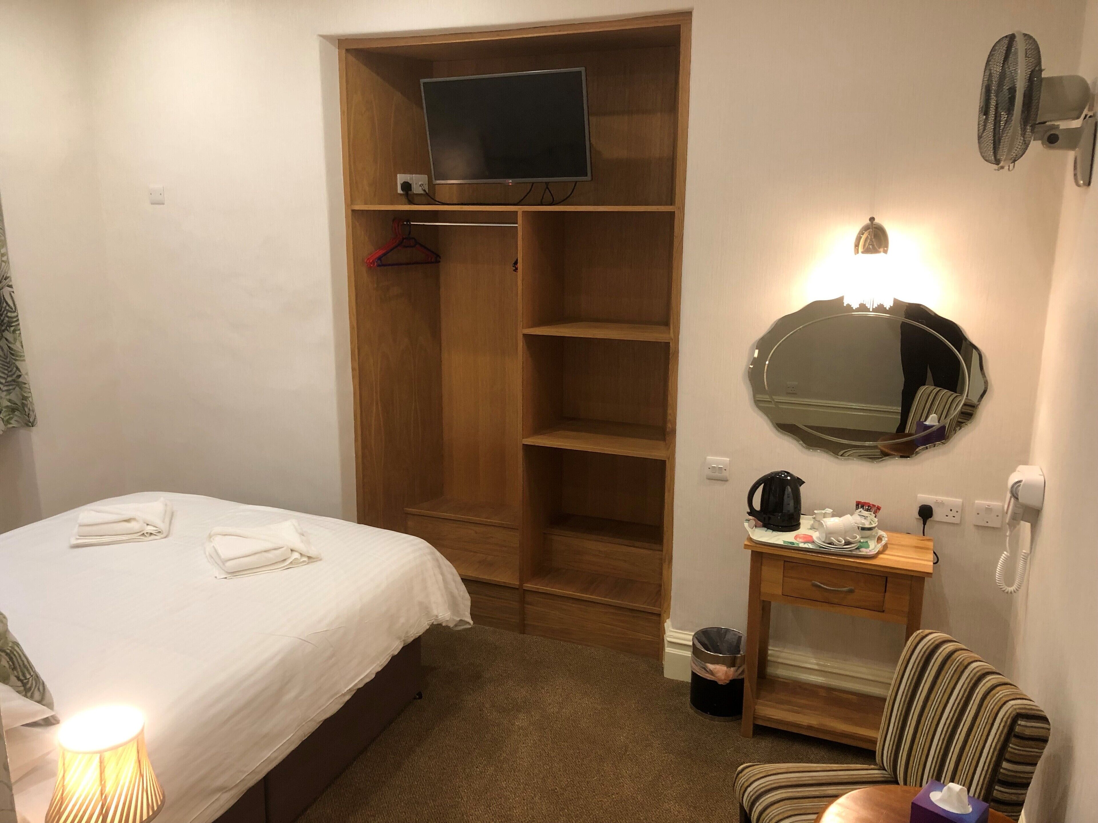 A hotel room with a bed, a wooden wardrobe, a television, a mirror, a small table with toiletries, and a chair.